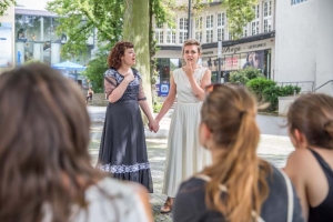 Zwei Frauen in Kleidern stehen vor dem Kino in Hildesheim und halten sich an den Händen. Die linke Frau scheint zu singen. Die Frauen werden von Publikum beobachtet.
