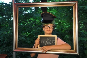 Ein Junge in Verkleidung schaut durch einen goldenen Bilderrahmen. Er hält eine kleine Tafel hoch. Darauf wurde mit Kreide geschrieben: "17/600". Hinter dem Jungen befinden sich Bäume.