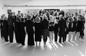 Viele Menschen verschiedener Altersklassen stehen in einem Kreis zusammen. Sie tragen dunkle Kleidung und sind barfuß. Das Foto ist schwarz-weiß.
