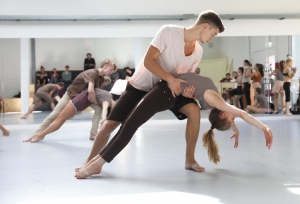 Junge Menschen tanzen paarweise in einem Tanzsaal. Die Männer halten die Frauen fest, die sich nach hinten lehnen. Im Hintergrund stehen viele Menschen und schauen zu.