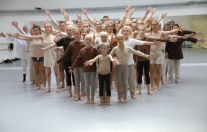 Kinder und Jugendliche stehen in einem Pulk zusammen und zeigen eine Tanzperformance. Sie breiten die Arme nach links und rechts aus. Alle tragen weiße oder braune Kleidung und sind barfuß.