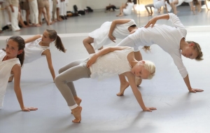 Kinder in weißer Kleidung zeigen eine Tanzperformance. Sie machen mit einem Arm eine Brücke. Den rechten Arm haben sie über ihren Bauch gelegt.