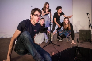 Ein junger Mann sitzt auf einer Bühne und blickt in die Kamera. Im Hintergrund stehen junge Frauen um einen Eimer herum an einem Mikrofon.