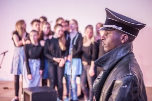 Eine farbige Person in SS-Uniform ist im Profil zu sehen. Im Hintergrund steht ein Pulk von jungen Menschen und schaut die Person an.