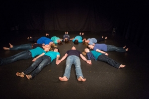 Menschen liegen im Kreis auf einer Bühne, mit den Köpfen nach innen. Sie halten sich an den Händen. Alle Menschen tragen blaue T-Shirts, Jeanshosen und sind barfuß.