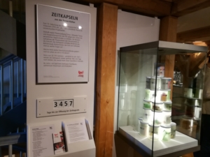 Eine Vitrine in einem Museum. In der Vitrine sind leuchtende Becher aufeinander gestapelt. Neben der Vitrine befindet sich ein eingerahmtes Schild mit einem Text. Über dem Text steht: "Zeitkapseln aus der freien Kultur". Unter dem Schild ist ein Zähler angebracht. Der Zähler zeigt die Zahl 3457. Darunter steht: "Tage bis zur Öffnung der Zeitkapseln".
