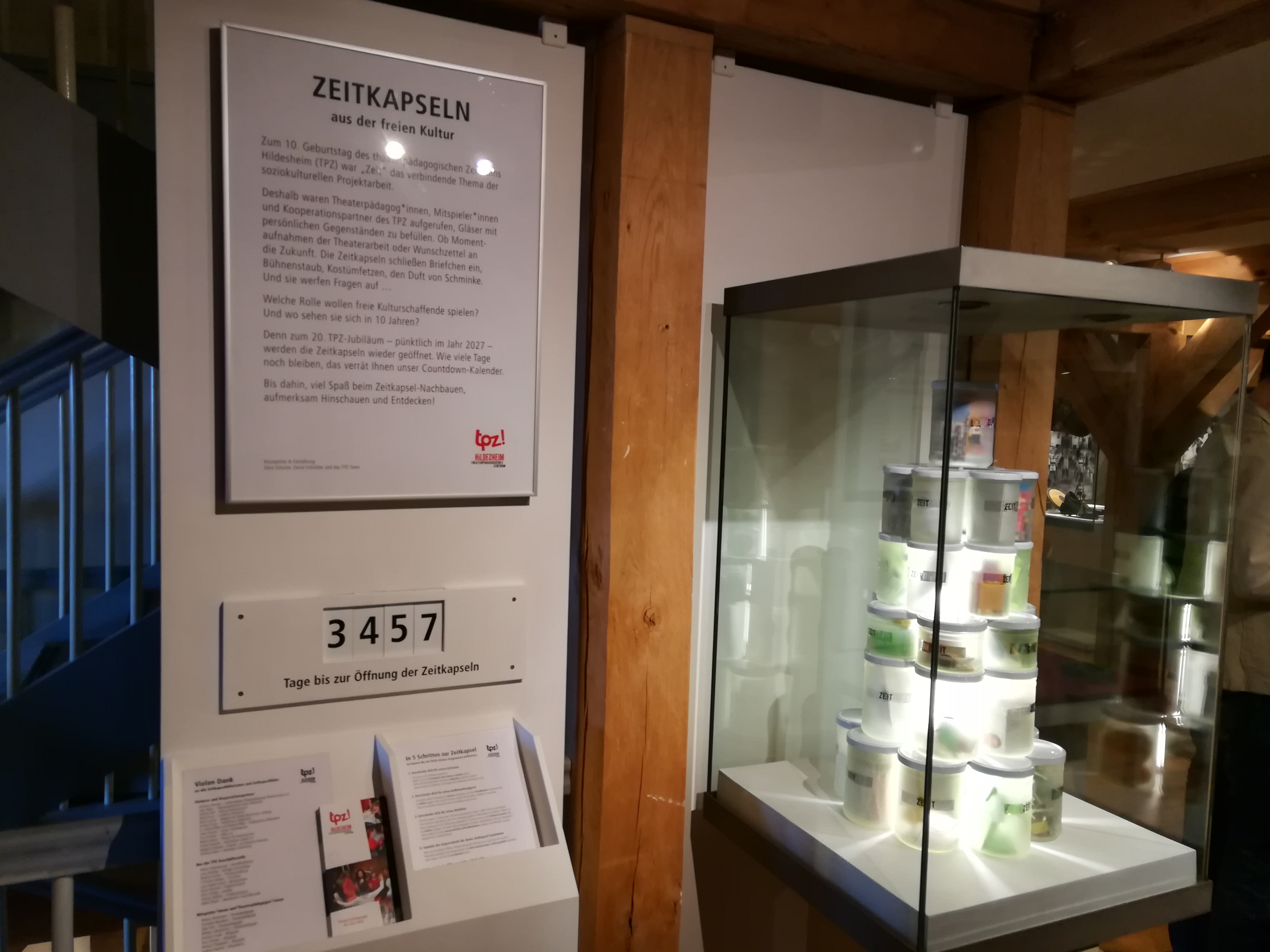 Eine Vitrine in einem Museum. In der Vitrine sind leuchtende Becher aufeinander gestapelt. Neben der Vitrine befindet sich ein eingerahmtes Schild mit einem Text. Über dem Text steht: "Zeitkapseln aus der freien Kultur". Unter dem Schild ist ein Zähler angebracht. Der Zähler zeigt die Zahl 3457. Darunter steht: "Tage bis zur Öffnung der Zeitkapseln".
