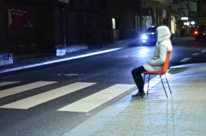 Eine Person sitzt auf einem orangenen Stuhl. Der Stuhl steht an einem Zebrastreifen an der Straße. Ein Auto nähert sich. Es ist dunkel.