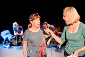 Das Bild entstand während einer Theateraufführung. Eine Frau im grünen T-Shirt hält einer Frau im grauen T-Shirt ein Mikrofon entgegen. Die Frau im grauen T-Shirt trägt eine Taucherbrille auf der Stirn und ein rotes Kletterseil in der Hand. Um ihren Hals hängt eine rote Trillerpfeife. Im Hintergrund hocken einige Menschen. Einer der Menschen hält eine weiße Stange zwischen den Händen. Es sieht aus, als wolle er damit Feuer machen. Ein anderer Mann hockt neben ihm und pustet ans Ende der Stange.