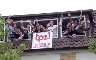 Viele Menschen winken aus drei Fenstern eines Dachgeschosses. Zwei Personen halten ein Banner in der Hand. Auf dem Banner ist das Logo vom TPZ abgebildet. Bei den Menschen handelt es sich um die Mitarbeiter und Mitarbeiterinnen des TPZ. Sie winken aus der Geschäftsstelle am Ratsbauhof.