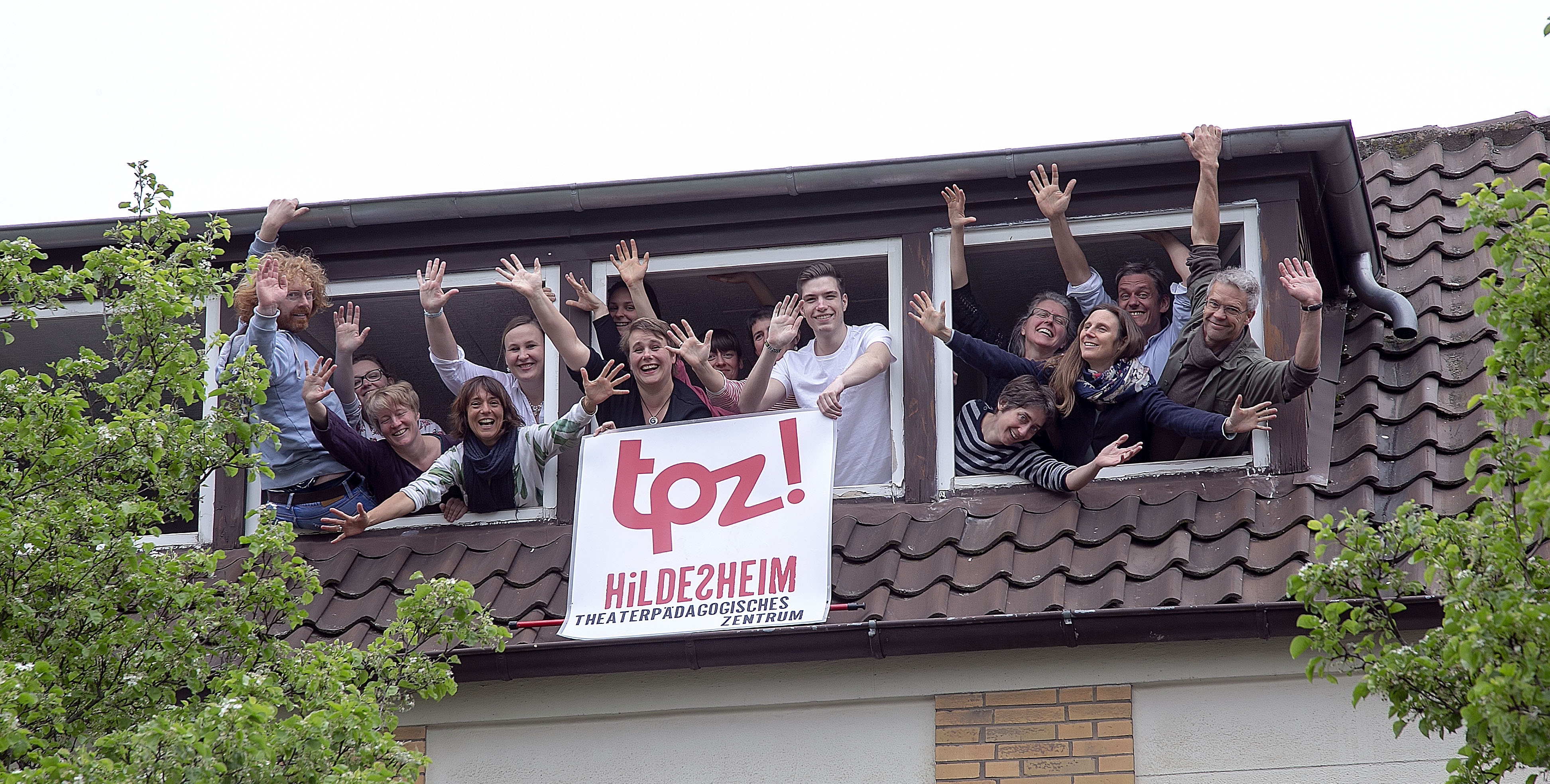 Viele Menschen winken aus drei Fenstern eines Dachgeschosses. Zwei Personen halten ein Banner in der Hand. Auf dem Banner ist das Logo vom TPZ abgebildet. Bei den Menschen handelt es sich um die Mitarbeiter und Mitarbeiterinnen des TPZ. Sie winken aus der Geschäftsstelle am Ratsbauhof.