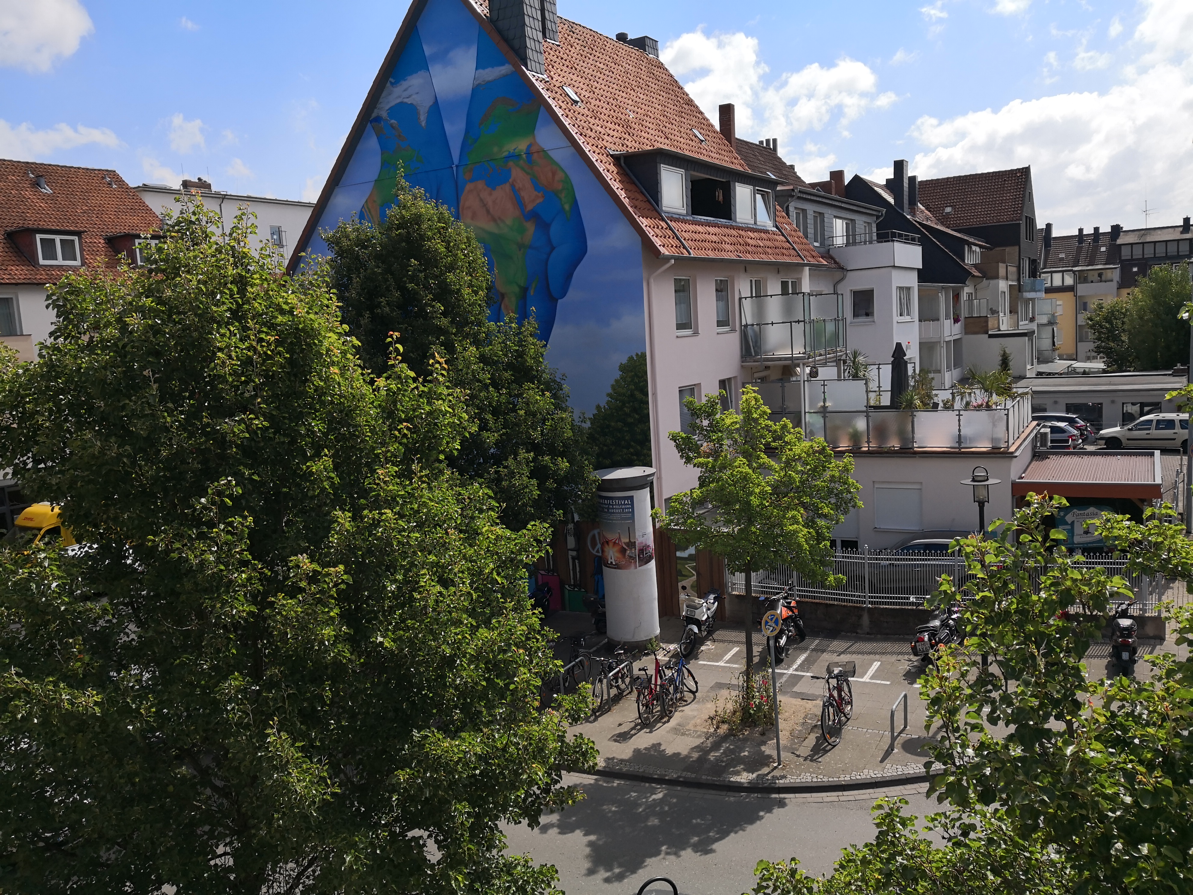 Der Ausblick aus der Geschäftsstelle des TPZ am Ratsbauhof. Man blickt auf grüne Bäume, Fahrräder, eine Litfaßsäule und einige Wohnhäuser. An einem der Wohnhäuser ist ein großes Bild gemalt. Das Bild zeigt zwei Hände, die wie eine Erde bemalt sind. Die Hände sind zu einer Schale geformt.
