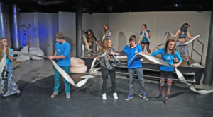 Das Bild ist bei einer Theateraufführung entstanden. Jugendliche stehen in zwei Reihen hintereinander. Die hintere Reihe steht erhöht auf einem Podest. Die Jugendlichen tragen blaue T-Shirts, auf denen ein Wasserhahn abgebildet ist. Beide Reihen halten gemeinsam jeweils einen Feuerwehrschlauch hoch. Der Schlauch ist leer.