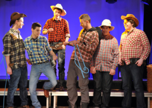 Berufsschüler*innen Das Foto entstand während einer Theaterraufführung. Jugendliche stehen auf einer Bühne. Sie tragen Hemden mit Karomuster und Cowboyhüte. Der Mann in der Mitte hält ein blaues Seil.
