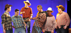 Das Foto entstand während einer Theaterraufführung. Jugendliche stehen auf einer Bühne. Sie tragen Hemden mit Karomuster und Cowboyhüte. Der Mann in der Mitte hält ein blaues Seil.