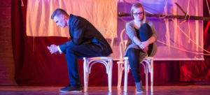 Das Foto entstand während einer Theateraufführung. Zwei Personen sitzen auf weißen Stühlen auf der Bühne. Sie schauen zu Boden. Die rechte Person hat eines ihrer Beine angewinkelt. Die linke Person hat sich abgewandt und sitzt seitlich auf dem Stuhl. Im Hintergrund hängen weiße Tücher.