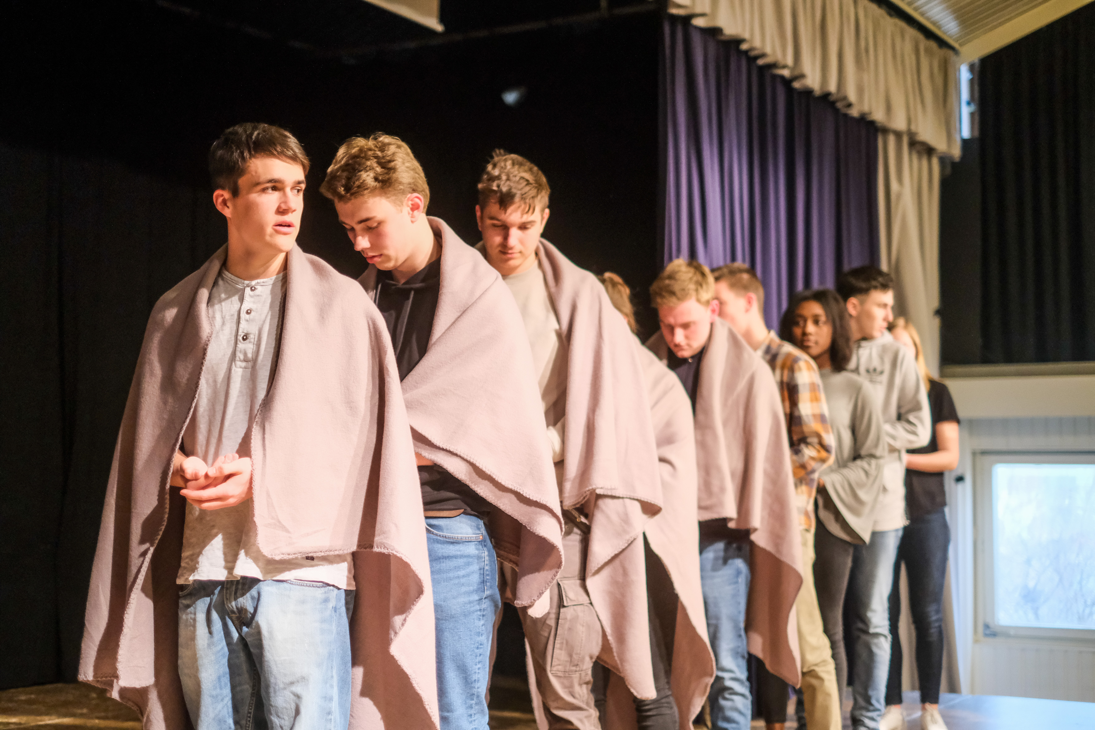 Das Foto entstand während einer Theaterprobe. Schüler und Schülerinnen stehen in einer Reihe. Um ihre Schultern hängen braune Decken. Die Schüler und Schülerinnen schauen mit ernstem Blick nach unten.