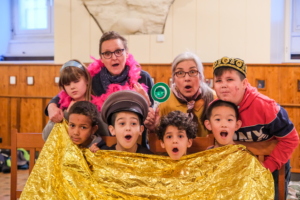 Das Foto entstand bei einer Theateraufführung. Acht Personen schauen mit offenem Mund in die Kamera. Die Personen sind in zwei Reihen aufgeteilt: Vorne sitzen vier Kinder auf Holzstühlen. Die Kinder haben eine goldene Rettungsdecke bis zum Kinn gezogen. Eines der Kinder trägt eine Polizeimütze. Hinter den vier Kindern stehen vier weitere Personen: Zwei Frauen in der Mitte und zwei Kinder links und rechts. Eine der Frauen trägt eine pinke Federboa um den Hals. Die andere Frau hält eine Polizeikelle hoch.
