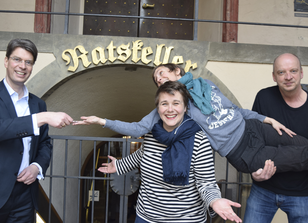 Vier Personen befinden sich vor dem Ratskeller in Hildesheim: Der Oberbürgermeister von Hildesheim, Ingo Meyer, die Geschäftsführerin des TPZ, Katrin Löwensprung, und zwei Theaterpädagogen und -pädagoginnen vom TPZ. Katrin Löwensprung und der Theaterpädagoge tragen die andere Theaterpädagogin. Der Bürgermeister trägt einen Anzug und übergibt einen Schlüssel an die drei Personen vom TPZ.