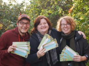 Drei Personen schauen lächelnd in die Kamera. Die Personen halten Flyer für ein Projekt vom TPZ hoch. Auf den Flyern steht: "Es war einmal in Eime". Die Personen stehen vor grünen Bäumen.