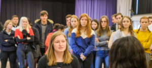 Zwei Frauen sitzen sich auf Stühlen gegenüber. Hinter der einen Frau stehen viele Jugendliche und schauen mit ernstem Blick nach vorne.