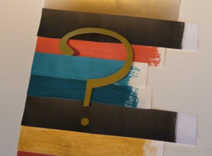 Bunt bemalte Streifen aus Papier liegen auf einem Tisch. Über den Streifen aus Papier liegt ein goldenes Fragezeichen.