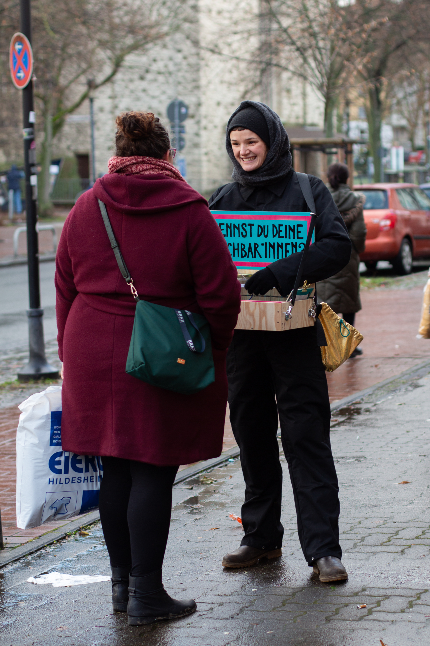 Eine Frau mit einem Bauchladen spricht mit einer anderen Frau. In dem Bauchladen ist ein Schild angebracht. Auf dem Schild steht: "Kennst du deine Nachbar*innen?"