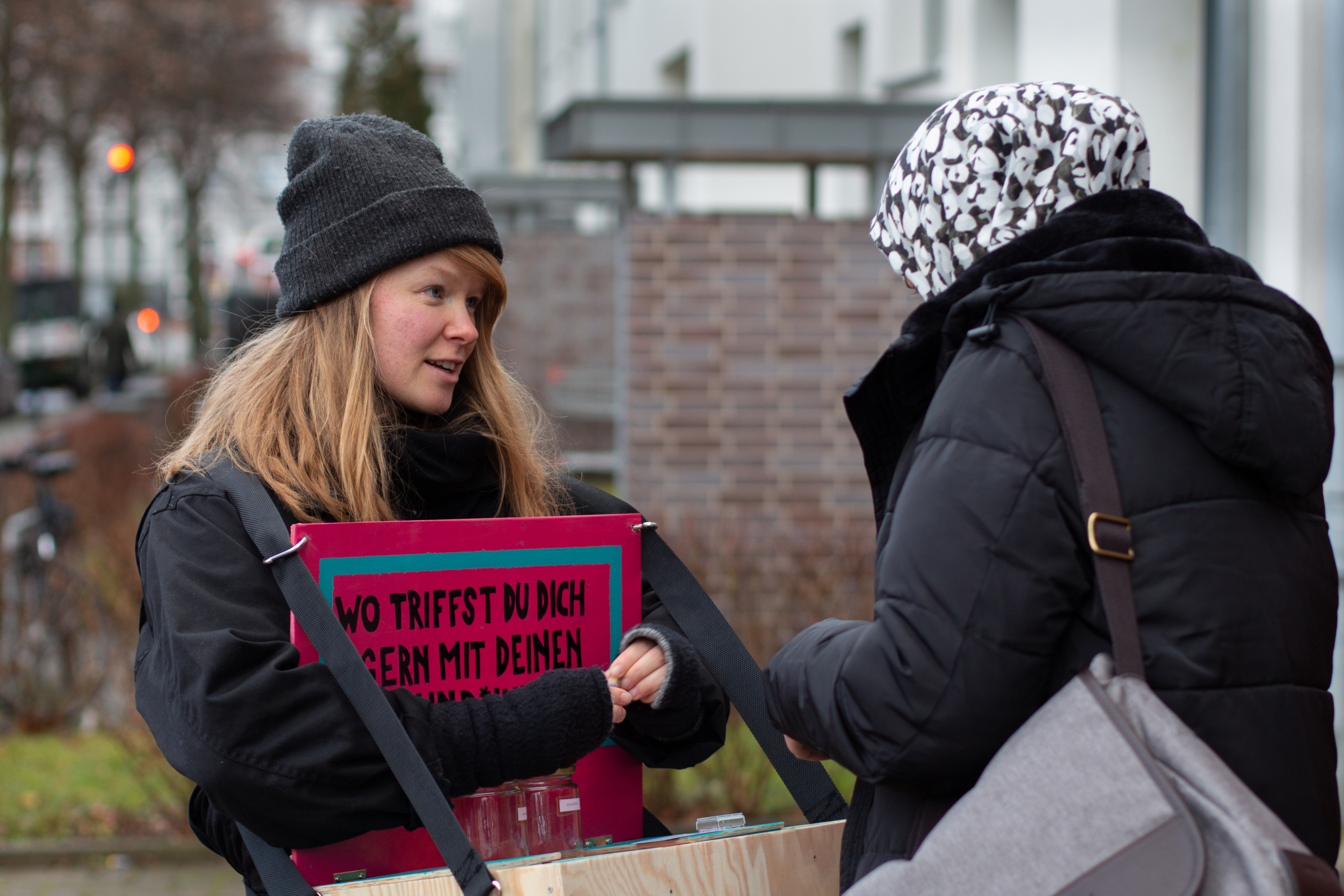 Eine Frau mit einem Bauchladen spricht mit einer Frau mit Kopftuch. Im Bauchladen ist ein Schild angebracht. Darauf steht: "Wo triffst du dich gern mit deinen Freund*innen?"