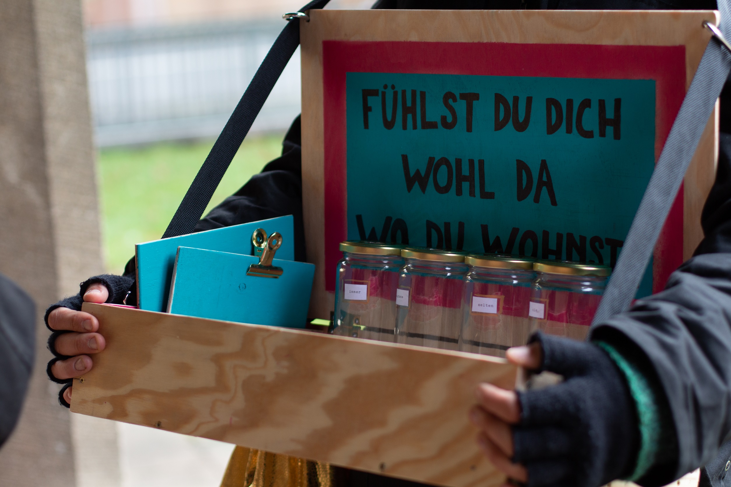 Eine Person mit einem Bauchladen. Im Bauchladen stehen leere Gläser. Über den Gläsern hängt ein Schild. Auf dem Schild steht: "Fühlst du dich wohl da wo du wohnst?"