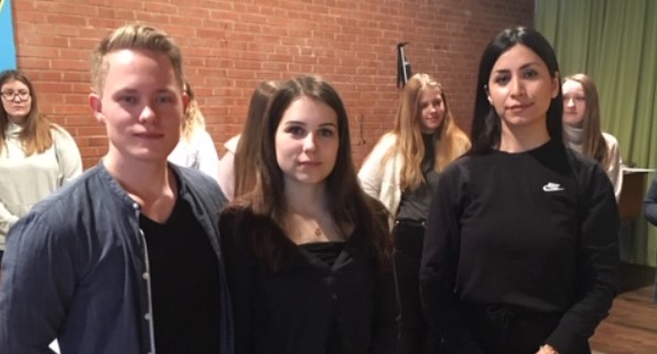 Drei junge Menschen blicken in die Kamera. Im Hintergrund stehen weitere junge Menschen.