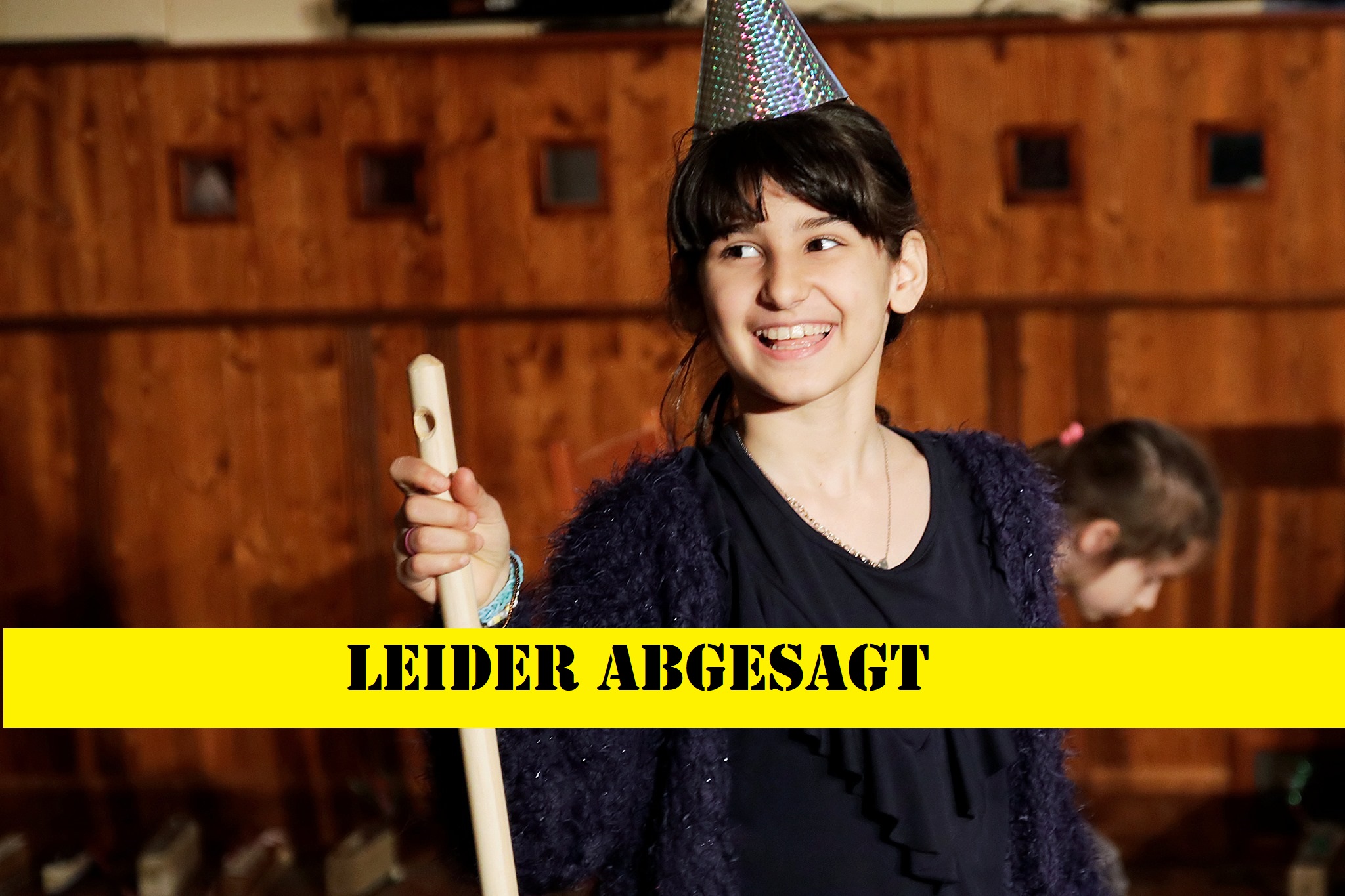 Ein Mädchen mit einem silbernen Hut auf dem Kopf. Das Mädchen hält eine Stange in der Hand. Über dem Bild ist ein gelber Schriftzug: "Leider abgesagt".