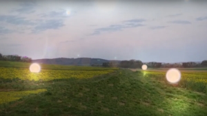 Ein grünes Feld unter grauem Himmel. Auf dem Feld sind mehrere Lichtpunkte zu sehen.