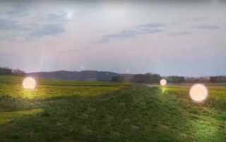Ein grünes Feld unter grauem Himmel. Auf dem Feld sind mehrere Lichtpunkte zu sehen.