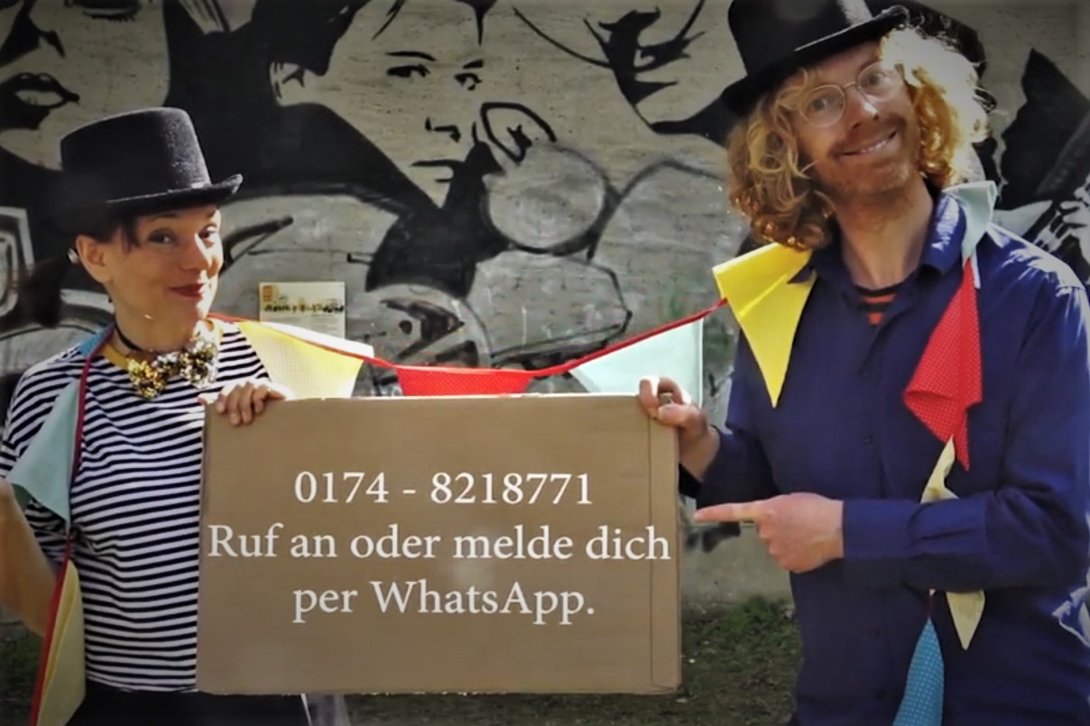 Zwei Menschen schauen in die Kamera. Sie sind als Zirkusdirektoren und Zirkusdirektorinnen verkleidet. Sie halten ein Schild hoch. Auf dem Schild steht: "0174 8218771 Ruf an oder melde dich per WhatsApp."