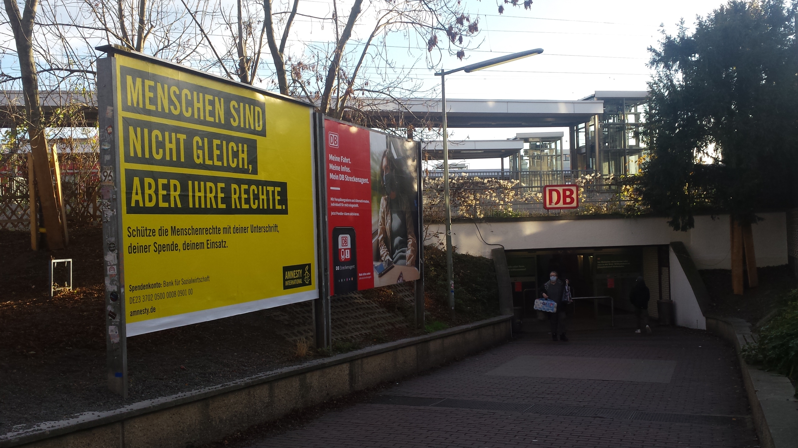 Der Hinterausgang von einem Bahnhof. Eine Person tritt aus dem Ausgang. Sie trägt eine Packung Toilettenpapier. Links im Bild befinden sich zwei Plakate. Die Plakate werben für "Amnesty International" und die Deutsche Bahn.