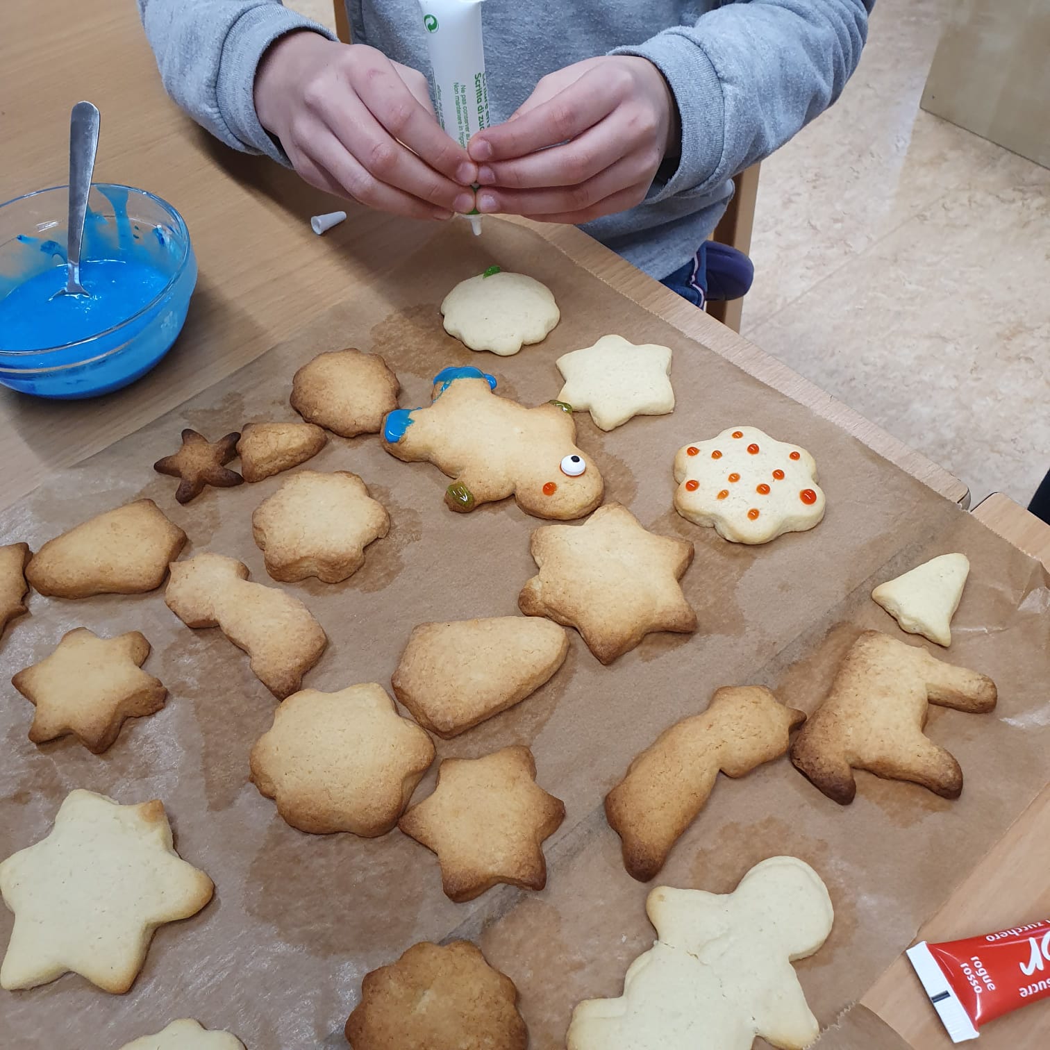 Plätzchen liegen auf einem Backpapier. Eine Person verziert die Plätzchen mit Lebensmittelfarbe.