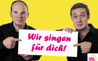 Zwei Männer halten ein Schild in die Kamera. Auf dem Schild steht: "Wir singen für dich!" Der Hintergrund des Bildes ist gelb.