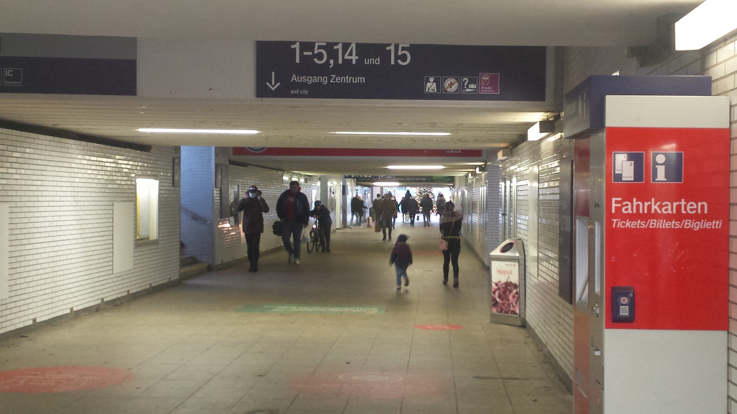 Die Unterführung in einem Bahnhof. Durch die Unterführung laufen viele Menschen. Rechts im Bild steht ein Fahrkartenautomat.