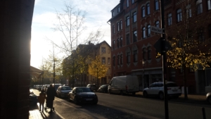 Eine Straße in Hildesheim. Links läuft eine Mutter mit ihrem Kind und einem Kinderwagen. Rechts befinden sich mehrere Altbauten. An der Straße parken Autos. Die Sonne scheint.