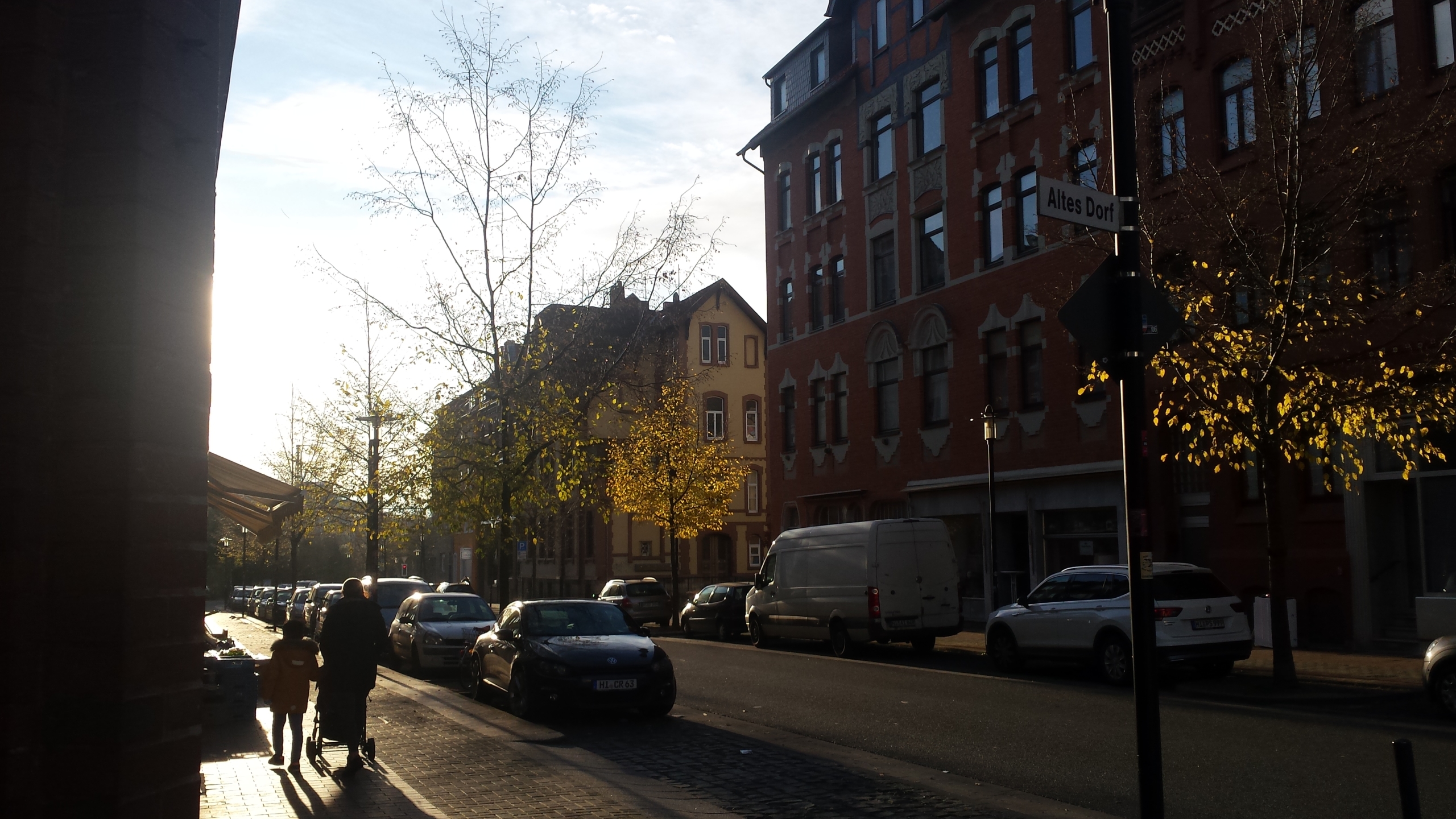 Eine Straße in Hildesheim. Links läuft eine Mutter mit ihrem Kind und einem Kinderwagen. Rechts befinden sich mehrere Altbauten. An der Straße parken Autos. Die Sonne scheint.