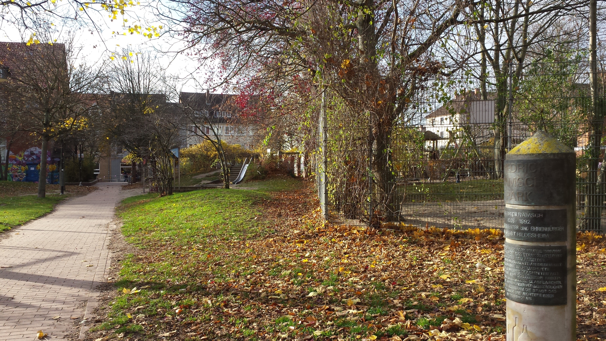 Ein Weg führt durch einen Park. Die Sonne scheint. Im Hintergrund sieht man einen Spielplatz.