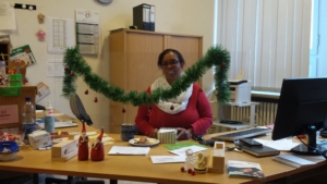 Eine Frau sitzt an einem Schreibtisch und lächelt in die Kamera. Auf dem Schreibtisch steht Weihnachtsdeko. Vor der Frau steht ein Teller mit Plätzchen.