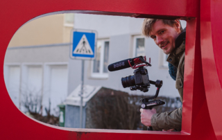 Ein Mann schaut in die Kamera. Er hält eine Kamera in der Hand. Er schaut durch ein rotes Gebilde.