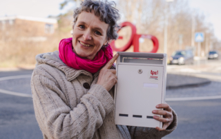 Eine Frau lächelt in die Kamera. Sie hält einen weißen Briefkasten in der Hand. Auf dem Briefkasten ist das Logo vom TPZ.