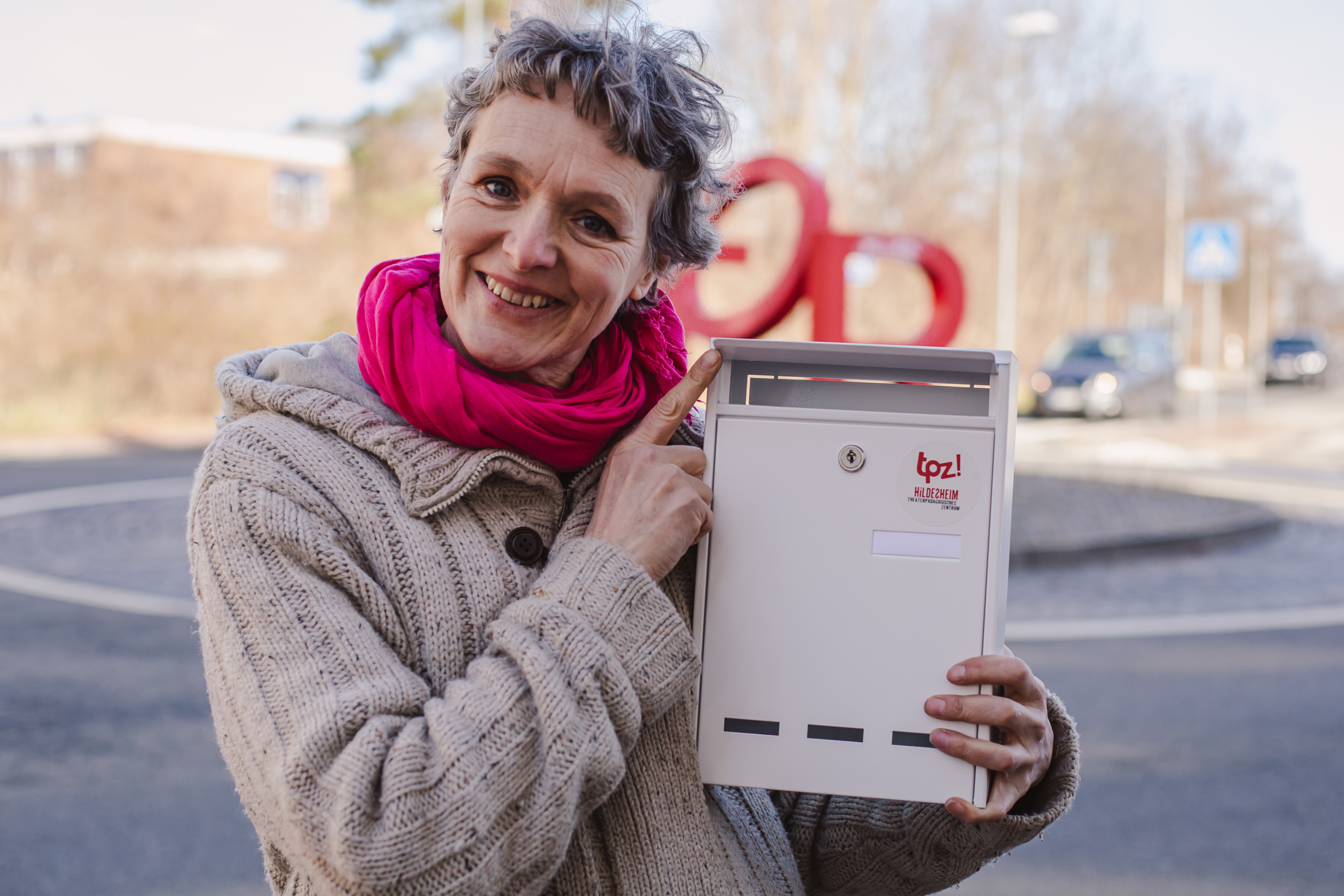 Eine Frau lächelt in die Kamera. Sie hält einen weißen Briefkasten in der Hand. Auf dem Briefkasten ist das Logo vom TPZ.