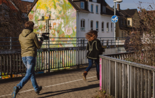 Zwei Personen rennen über eine Brücke. Die hintere Person trägt eine Kamera.