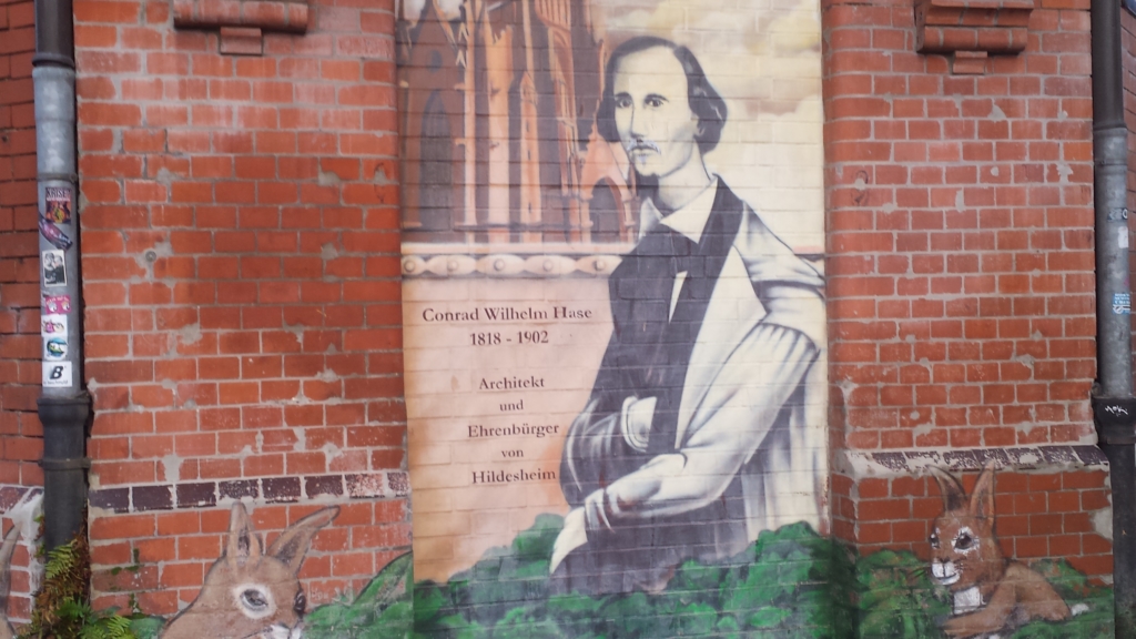 Eine Mauer, auf die ein Bild gemalt wurde. Das Bild zeigt einen Mann in einem Sakko. Neben dem Mann steht: "Conrad Wilhelm Hase 1818-1902, Architekt und Ehrenbürger von Hildesheim". Links und rechts von dem Mann sitzen zwei gemalte Hasen.
