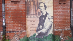 Eine Mauer, auf die ein Bild gemalt wurde. Das Bild zeigt einen Mann in einem Sakko. Neben dem Mann steht: "Conrad Wilhelm Hase 1818-1902, Architekt und Ehrenbürger von Hildesheim". Links und rechts von dem Mann sitzen zwei gemalte Hasen.