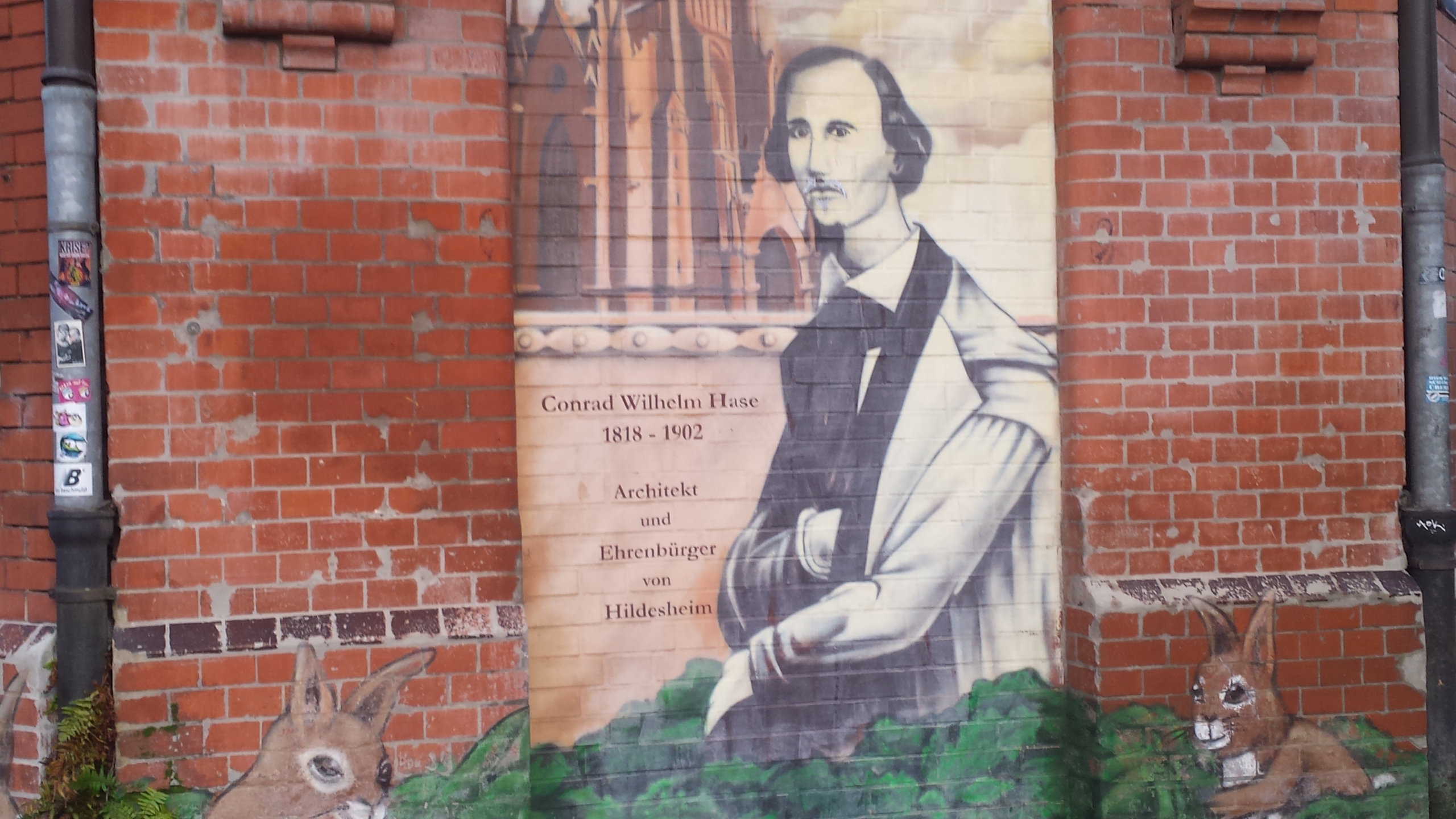 Eine Mauer, auf die ein Bild gemalt wurde. Das Bild zeigt einen Mann in einem Sakko. Neben dem Mann steht: "Conrad Wilhelm Hase 1818-1902, Architekt und Ehrenbürger von Hildesheim". Links und rechts von dem Mann sitzen zwei gemalte Hasen.