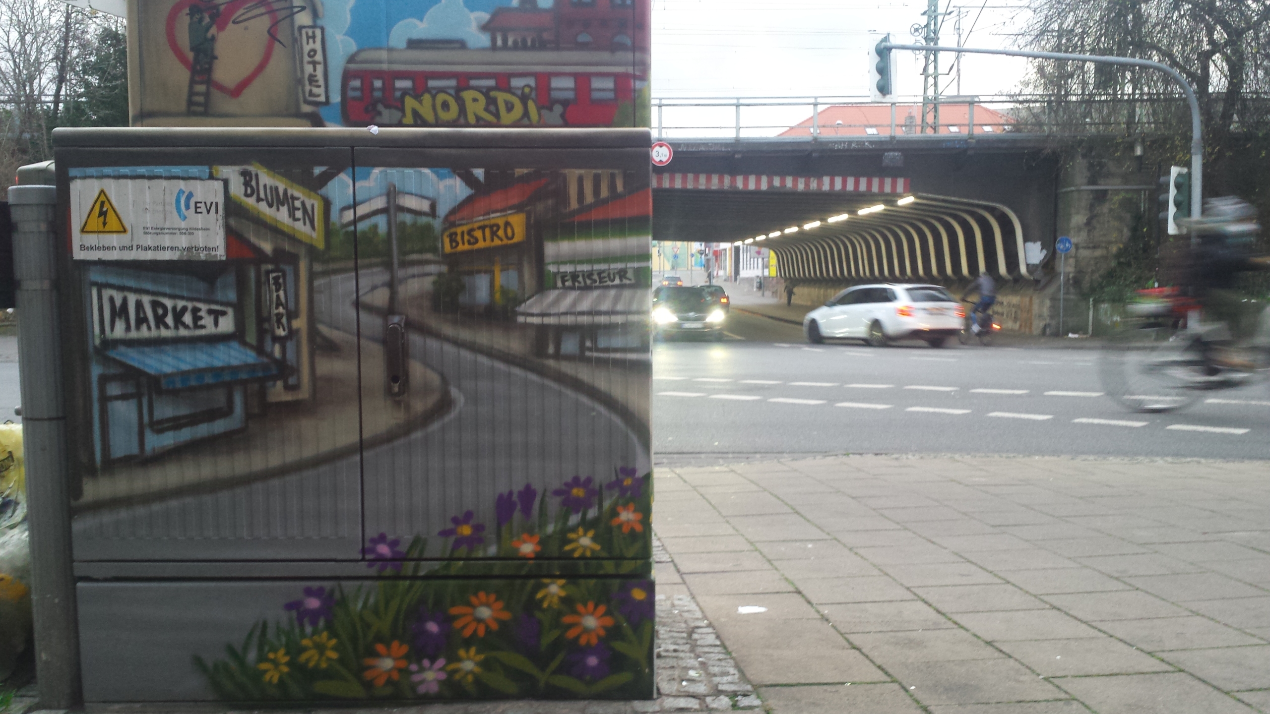 Ein Platz in Hildesheim. Im Vordergrund sieht man einen bunt bemalten Stromkasten. Im Hintergrund ist eine Unterführung zu sehen. Autos fahren auf einer Straße.